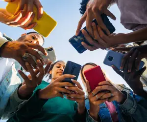 A low-angle shot of a diverse group of young adults collectively looking down at their smartphones. This visualizes the mobile-first audience captured through direct mobile lock screen communication and enterprise loyalty marketing initiatives.